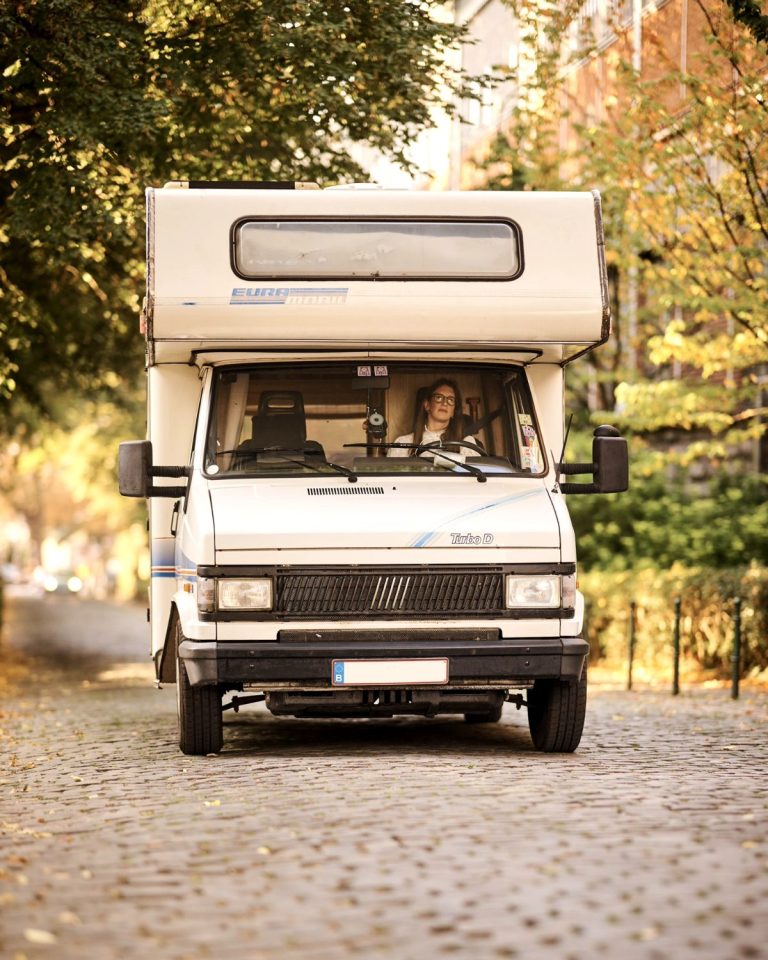 Emilie au volant de Gaston, son camping-car Fiat Ducato de 1991, sur une rue pavée bordée d'arbres